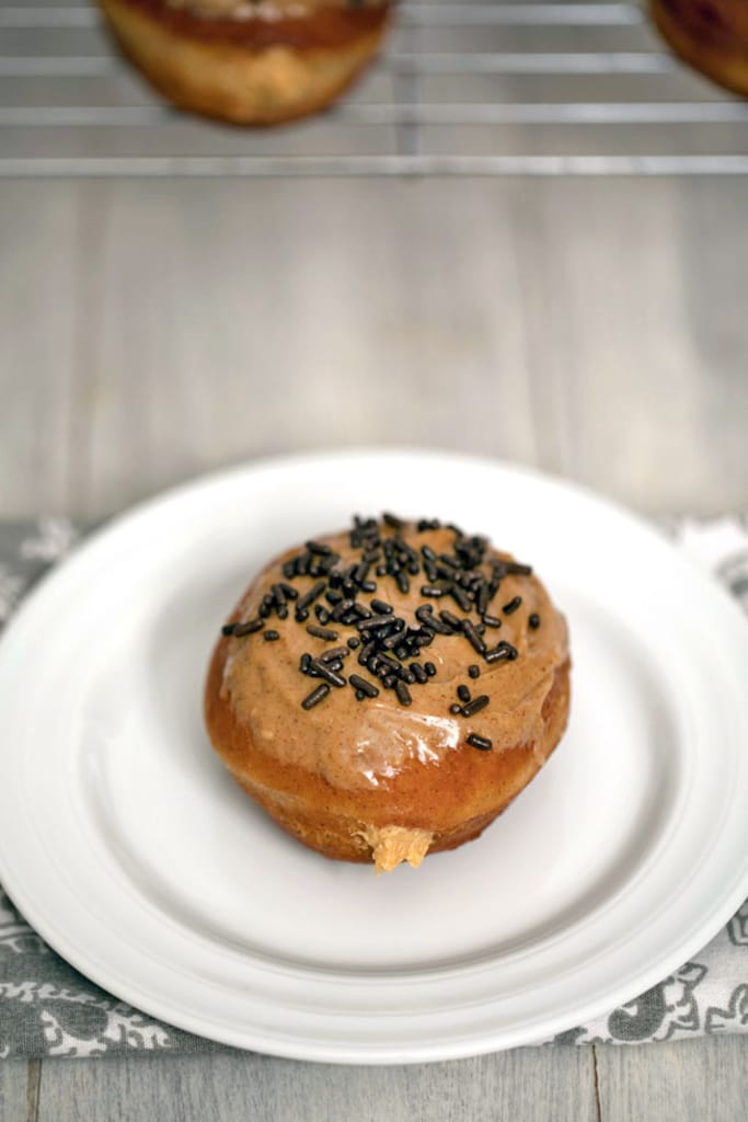 Overhead view of a cinnamon doughnut on a white plate and topped with peanut butter glaze and chocolate sprinkles with peanut butter cream filling peeping out.