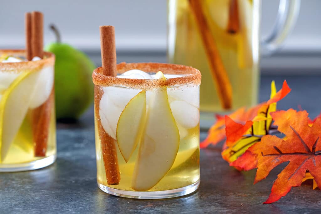 Landscape view of a glass of cinnamon pear sangria with second glass, pear, fall leaves, and pitcher of sangria in background