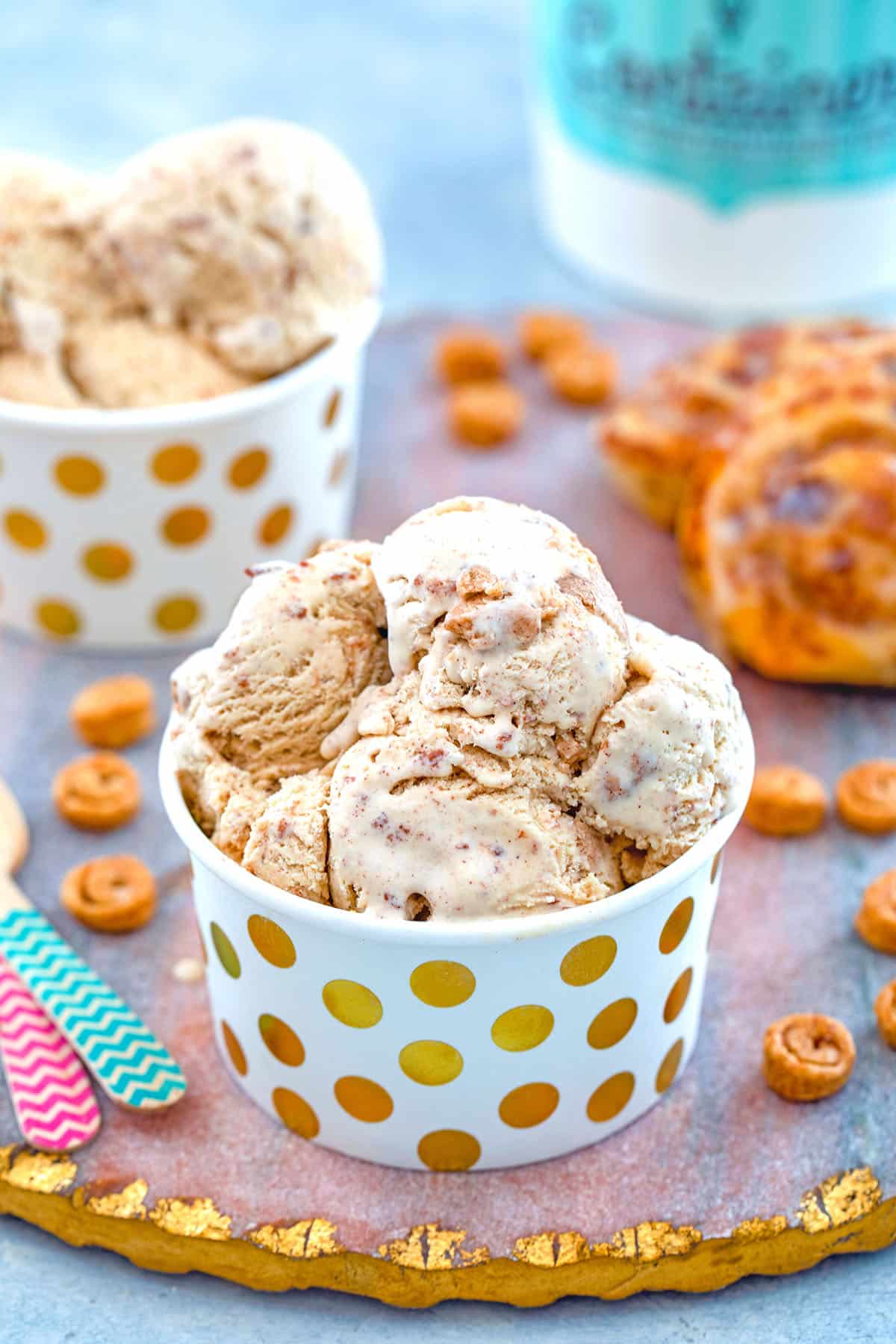 View of a small cup of cinnamon roll ice cream with mini cinnamon rolls, large cinnamon rolls, second cup of ice cream, and ice cream quart container in background with recipe title at top.