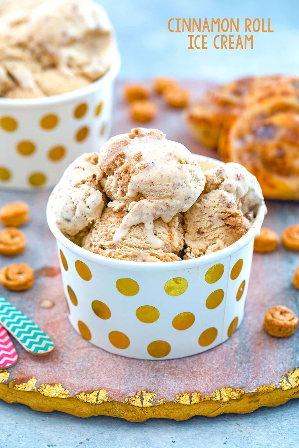 Head-on closeup view of a small cup of cinnamon roll ice cream with mini cinnamon rolls, large cinnamon rolls, and second cup of ice cream in background with recipe title at top.