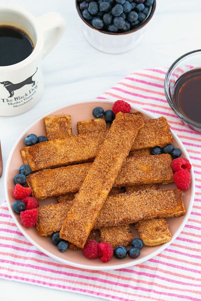 Cinnamon sugar frech toast sticks on a plate surrounded with berries with coffee in background.