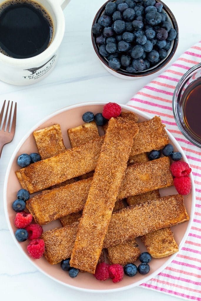 Overhead view of cinnamon sugar naan french toast sticks surrounded by blueberries and raspberries with bowl of blueberries and cup of coffee in background.