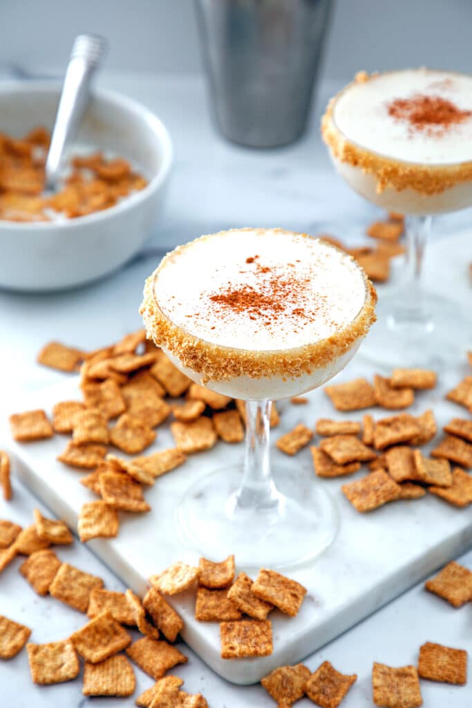 Overhead view of two Cinnamon Toast Crunch cocktails with cereal all around and bowl of cereal and cocktail shaker in background