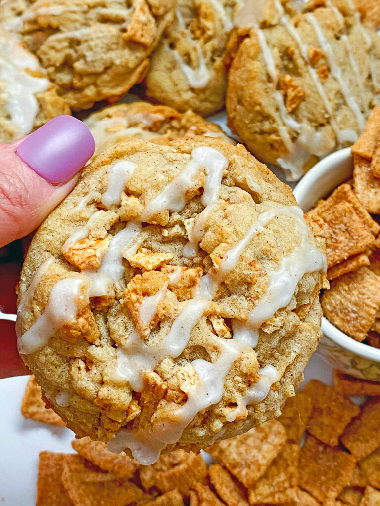 Close-up view of a hand holding a Cinnamon Toast Crunch cookie with cereal in the background.