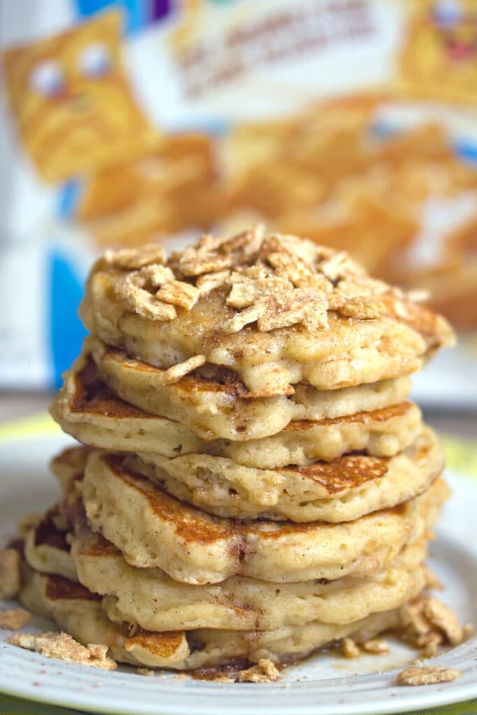 Head-on view of a big stack of cinnamon toast crunch pancakes with crushed Cinnamon Toast Crunch cereal on top and cereal box in the background