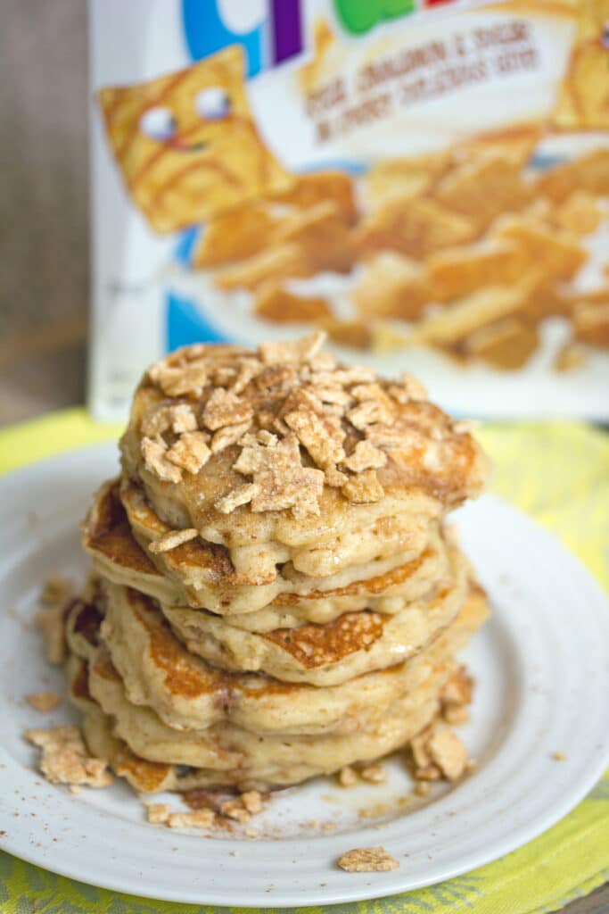 Overhead view of a stack of Cinnamon Toast Crunch pancakes with cereal crushed over the top on a white plate with cereal box in the background