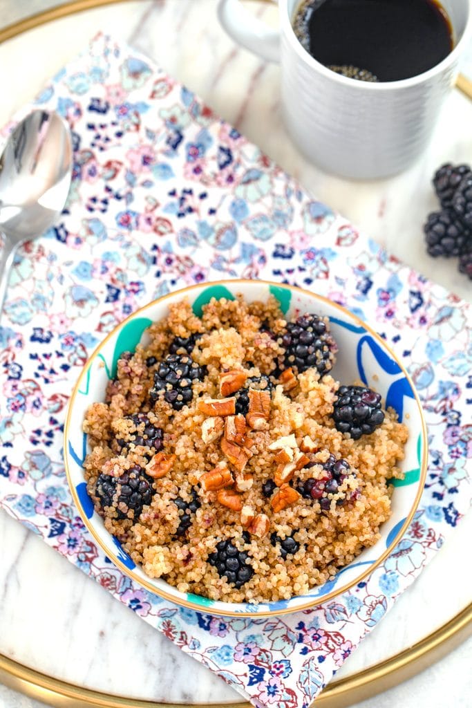 Cinnamon and Blackberry Breakfast Quinoa -- Quinoa for breakfast? It's not as weird as you think! This Cinnamon and Blackberry Breakfast Quinoa will keep you full and satisfied all morning long. Not to mention, it's a delicious way to start your day | wearenotmartha.com Overhead view of bowl of cinnamon and blackberry breakfast quinoa topped with pecans on a flowered towel on marble surface with cup of coffee and spoon in the background