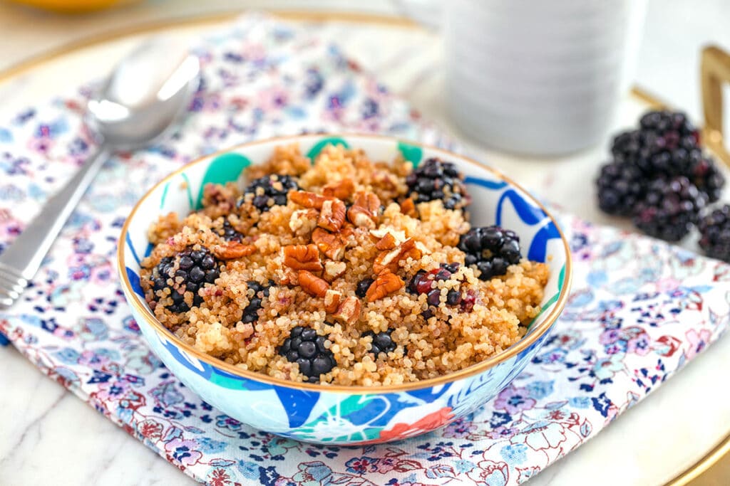 Landscape photo of colorful bowl of cinnamon and blackberry breakfast quinoa topped with pecans on flowered towel with spoon, blackberries, and mug in the background