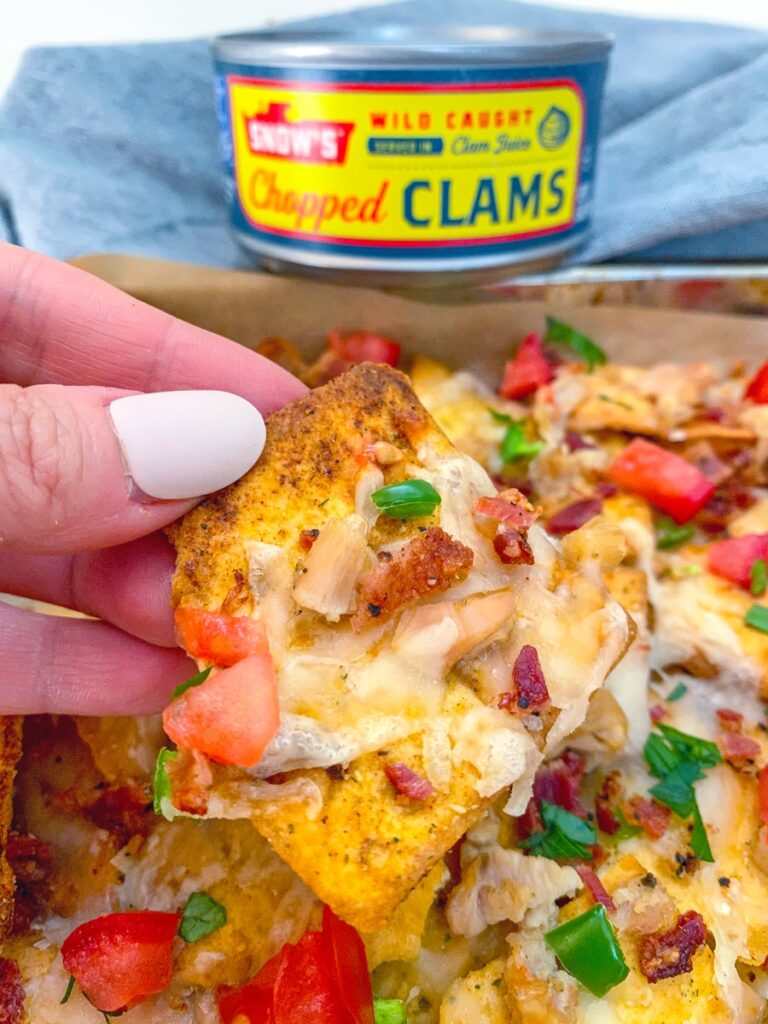 Hand holding up a clam nacho with can of chopped clams in the background