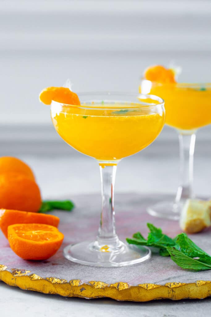Head-on view of two clementine ginger cocktails surrounded by clementines, mint leaves, and a piece of ginger