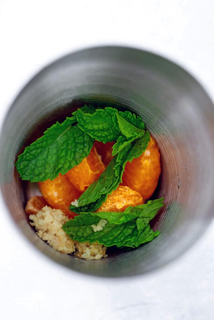 Overhead view of clementine wedges, mint leaves, and grated ginger in cocktail shaker
