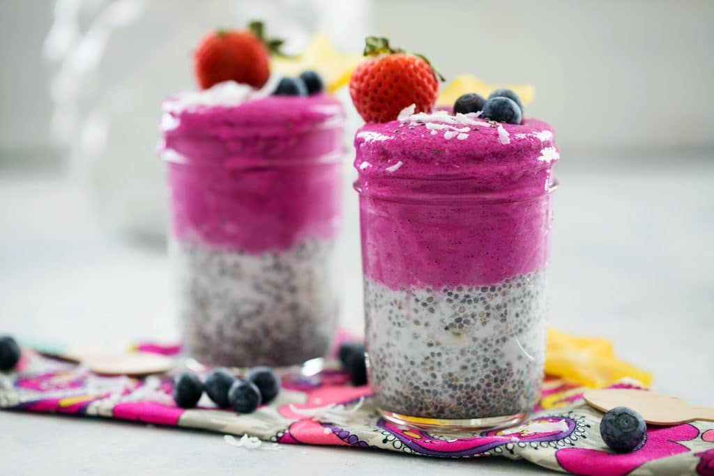 Landscape view of two coconut chia dragon fruit smoothie jars topped with coconut and fruit and surrounded by blueberries and shredded coconut