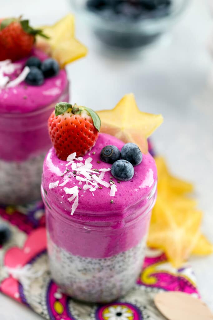 Overhead view of a bright pink coconut chia dragon fruit smoothie jar topped with shredded coconut, blueberries, a strawberry, and a slice of star fruit with second jar in background