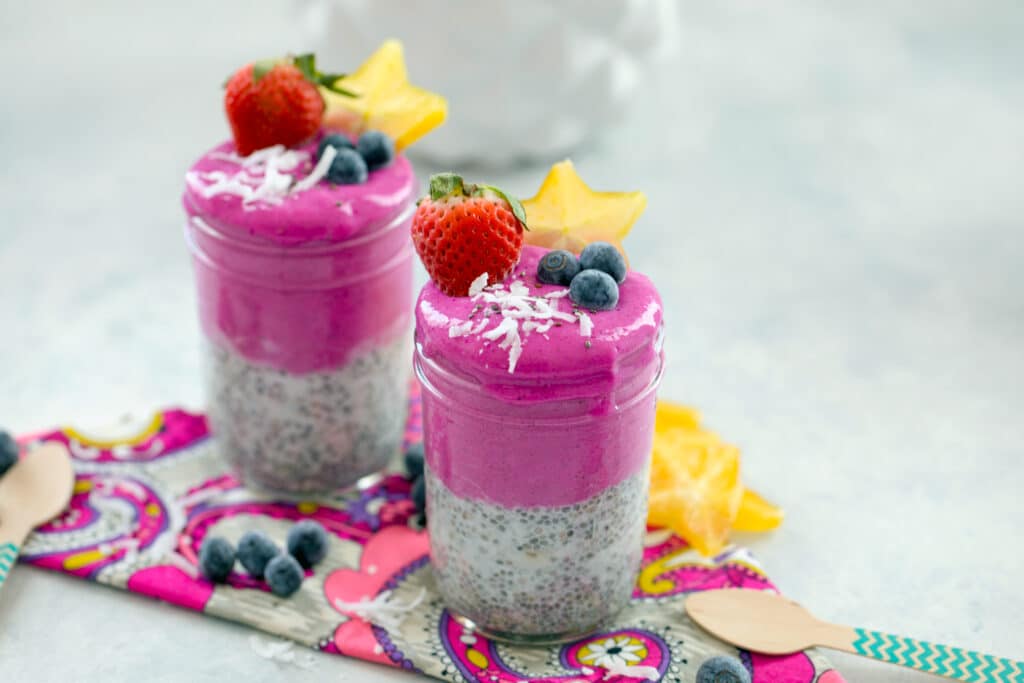 Landscape view of two coconut chia dragon fruit smoothie jars topped with coconut and fruit and surrounded by blueberries and shredded coconut