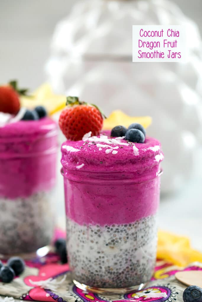 Head-on view of a bright pink coconut chia dragon fruit smoothie jar topped with shredded coconut, blueberries, strawberry, and star fruit with second smoothie jar in background and recipe title at top