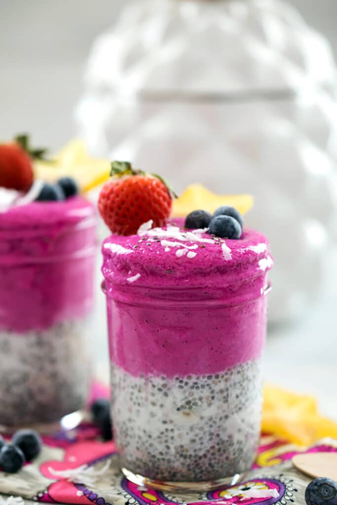 Head-on view of a bright pink coconut chia dragon fruit smoothie jar topped with shredded coconut, blueberries, strawberry, and star fruit with second smoothie jar in background