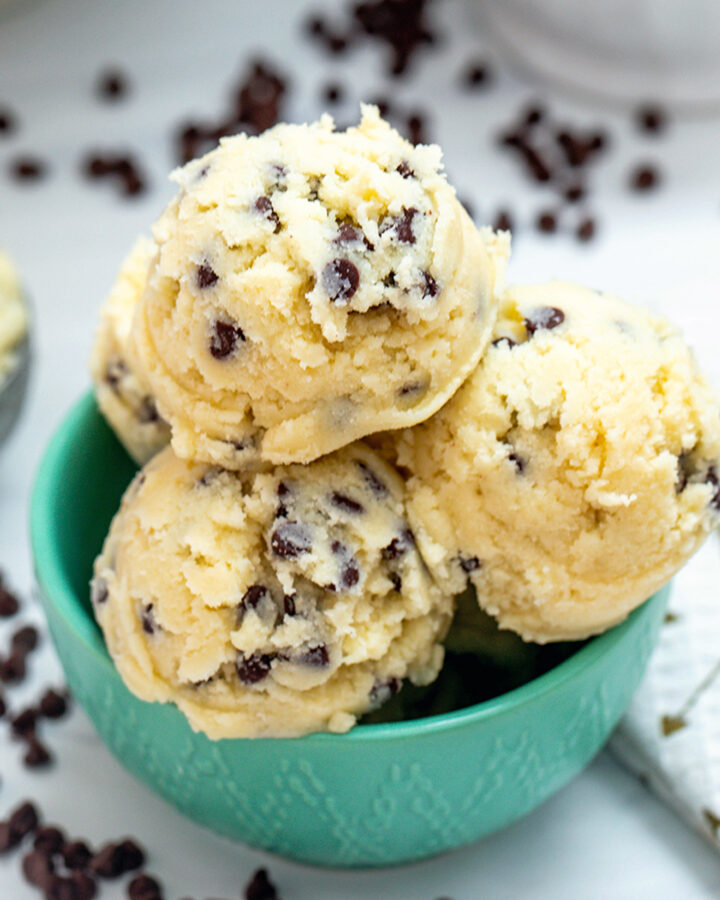 Overhead closeup view of a bowl of coconut flour cookie dough balls with chocolate chips, flour, and a scoop of cookie dough in background
