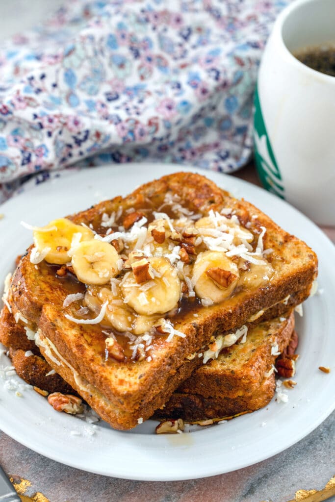 Head-on view of a stack of whole wheat coconut french toast topped with banana pecan caramel sauce and shredded coconut with cup of coffee in the background