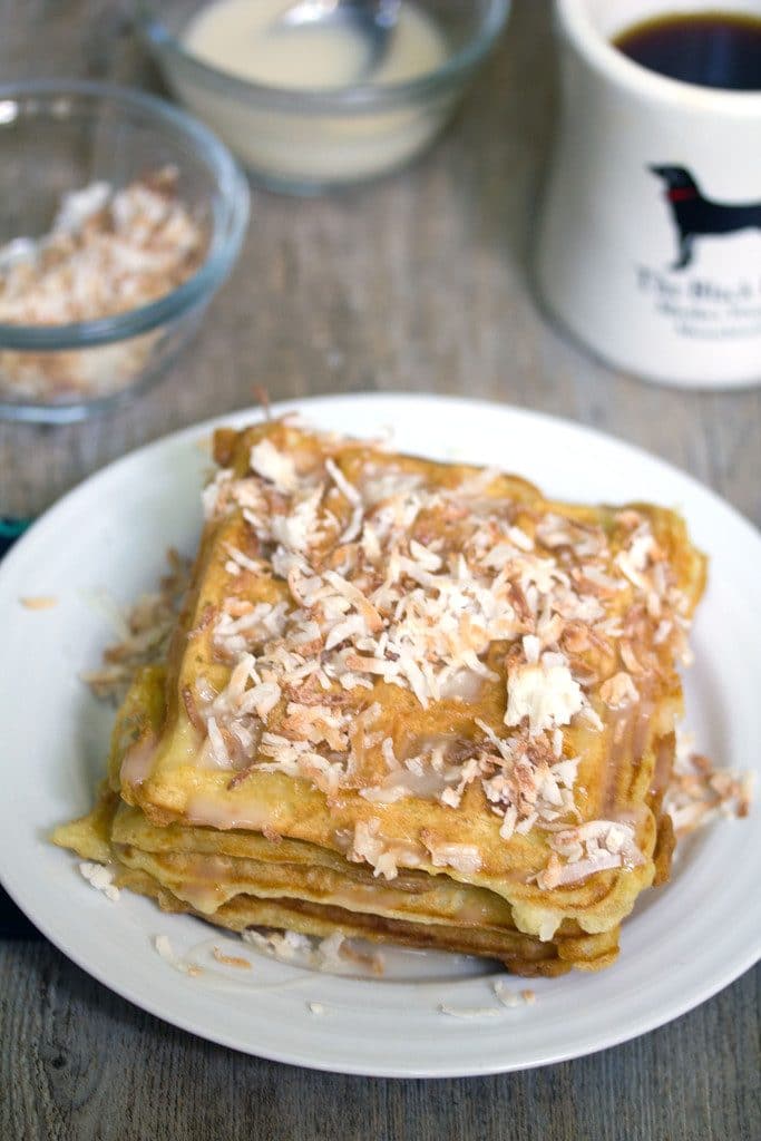 Overhead view of coconut waffles with vanilla coconut glaze topped with toasted coconut with coffee cup and bowls of toasted coconut and glaze in the background