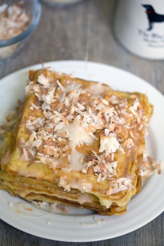 Closeup of stack of coconut waffles with vanilla coconut glaze and toasted coconut on a white plate