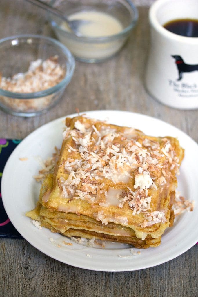 Overhead view of coconut waffles with vanilla coconut glaze topped with toasted coconut with coffee cup and bowls of toasted coconut and glaze in the background