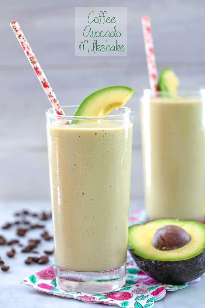 Head-on view of coffee avocado milkshake in a tall glass on a pink flower napkin with pink straw and avocado garnish and coffee beans, avocado half, and another drink in the background and "Coffee Avocado Milkshake" text