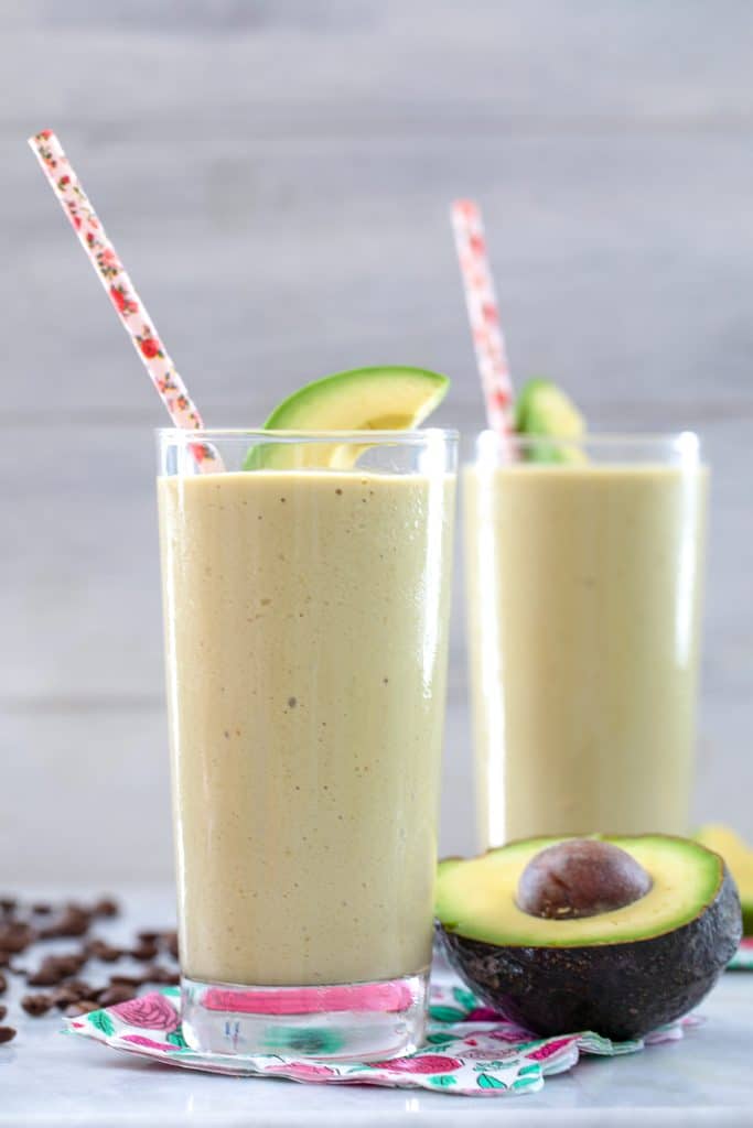 Up close head-on view of coffee avocado milkshake in a tall glass with pink straw and avocado garnish and another glass, avocado half, and coffee beans in the background