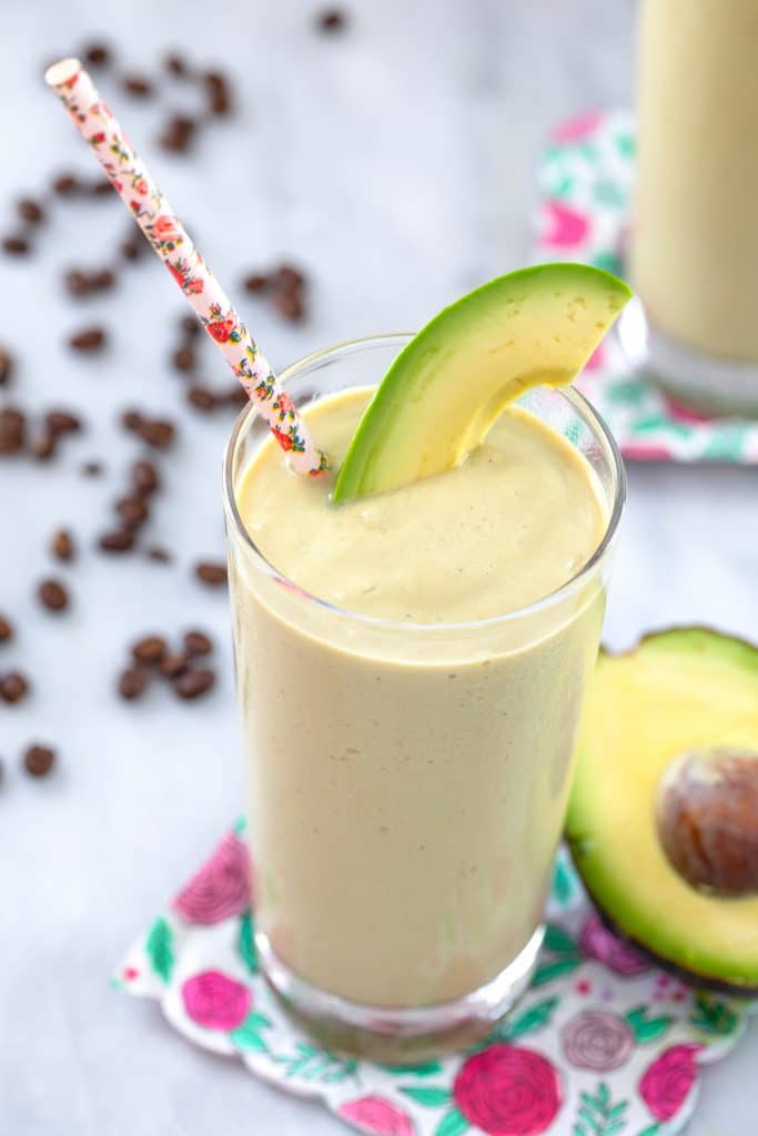 Overhead view of coffee avocado smoothie in tall glass with pink flower straw and avocado garnish on a pink flower napkin with avocado half and coffee beans in the background