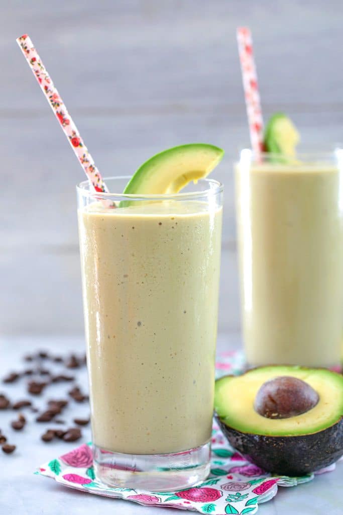 Head-on view of coffee avocado milkshake in a tall glass on a pink flower napkin with pink straw and avocado garnish and coffee beans, avocado half, and another drink in the background