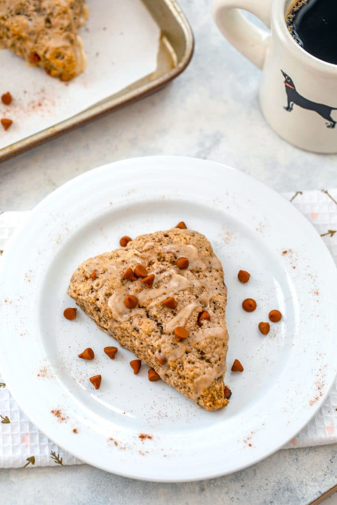 Overhead view of a coffee cinnamon scone on a white plate with cinnamon chips scattered around and a baking sheet with additional scones and cup of coffee in the background