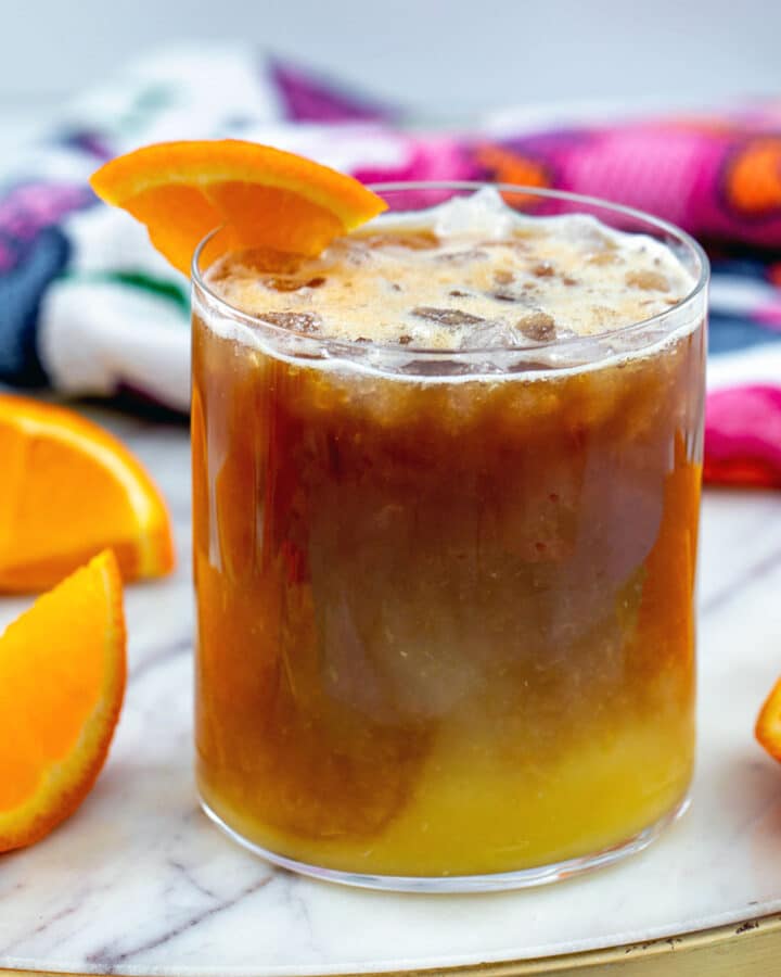 Closeup head-on view of coffee and orange juice in a glass with orange slices all around.