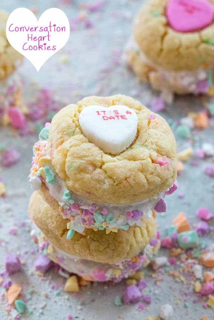 Head-on view of two conversation heart sandwich cookies with marshmallow fluff stacked on each other and surrounded by lots of crushed conversation hearts with recipe title at top