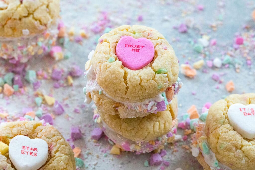 Landscape view of conversation heart sandwich cookies with marshmallow fluff surrounded by more conversation heart cookies and crushed conversation hearts.