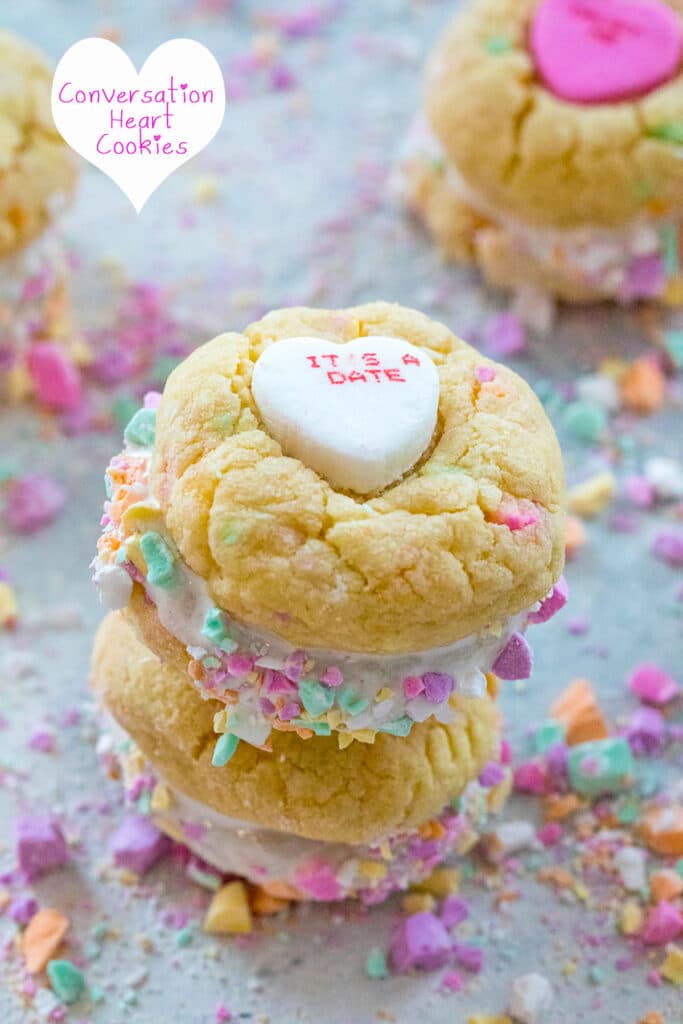 Head-on view of two conversation heart sandwich cookies with marshmallow fluff stacked on each other and surrounded by lots of crushed conversation hearts with recipe title at top.