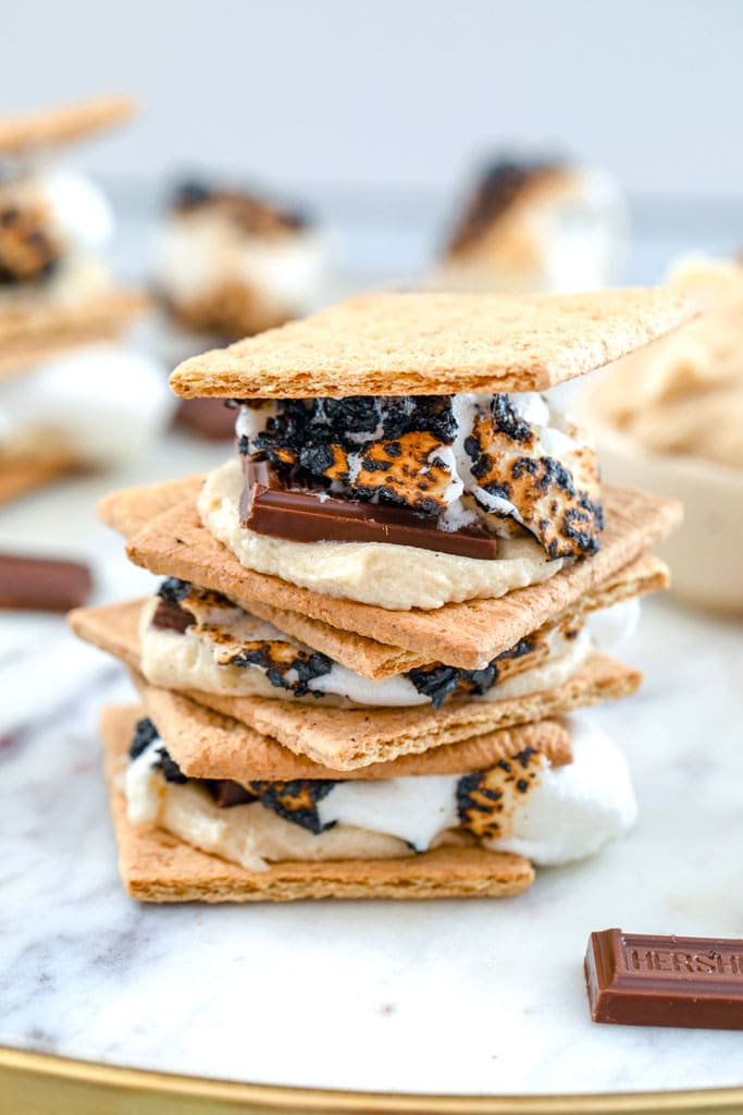 Head-on view of three cookie dough s'mores stacked on top of each other on a marble tray with chocolate bars and more s'mores in the background