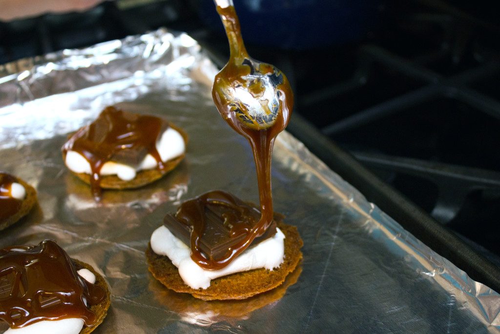 View of graham cracker cookies with marshmallows melted on top with a chocolate bar and salted caramel being drizzled over them