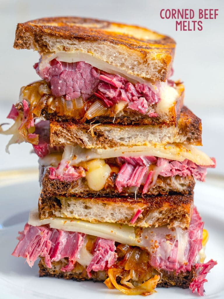 Head-on view of three halves of corned beef melts sandwiches stacked on top of each other on a white plate with the recipe title at top