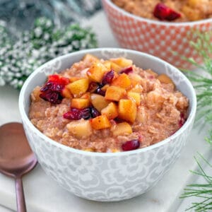 Head-on closeup view of a bowl of cranberry apple oatmeal with second bowl and holiday garland in background and recipe title at top