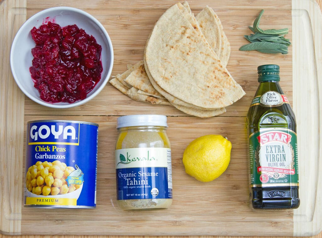 Overhead view of all ingredients for cranberry hummus and pita chips, including bowl of cranberry sauce, chick peas, tahini, lemon , olive oil, pita bread, and sage