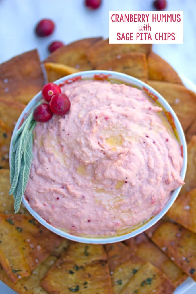 Overhead view of a bowl of cranberry hummus with cranberry and sage garnish and surrounded by sage pita chips, with "cranberry hummus" text at top of photo and "with sage pita chips" text at bottom