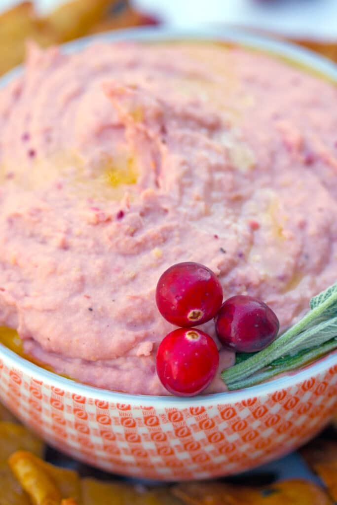 Close-up overhead view of red and white bowl filled with pink cranberry hummus with cranberry and sage garnish