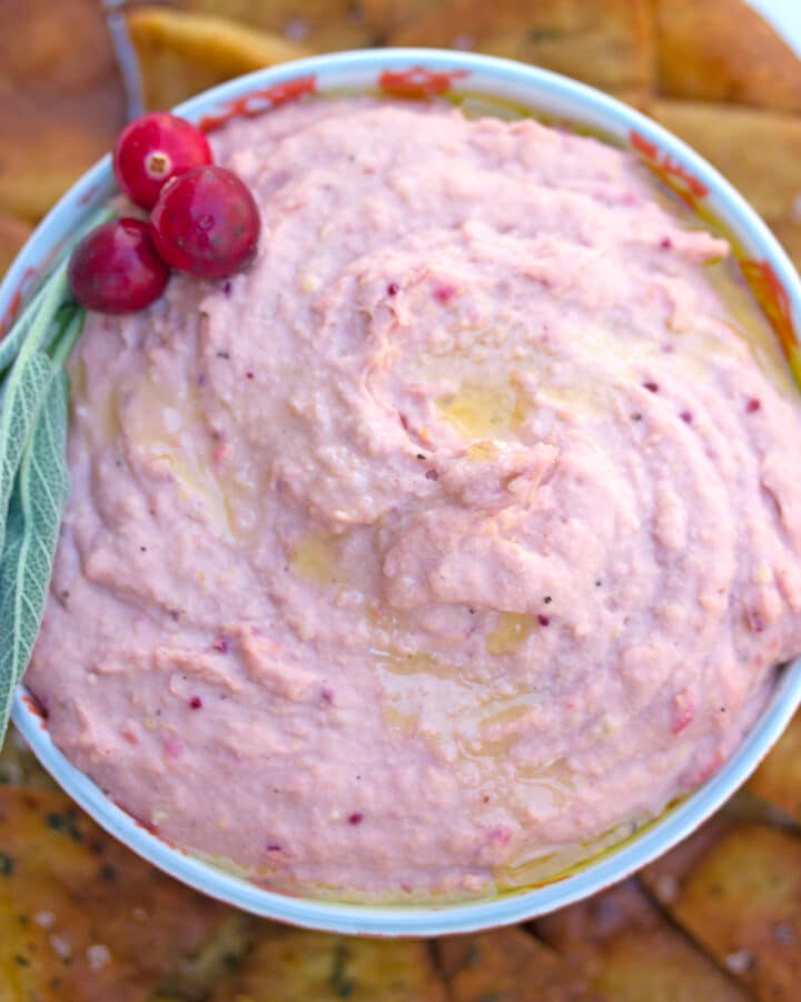 Overhead view of a bowl of cranberry hummus with cranberry and sage garnish and surrounded by sage pita chips