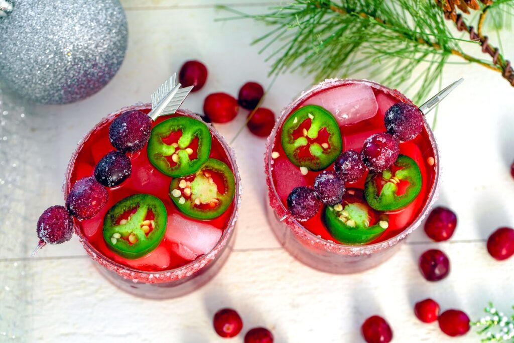 Bird's eye view of two cranberry jalapeño margaritas with cranberries, holly, and glitter ornament in background