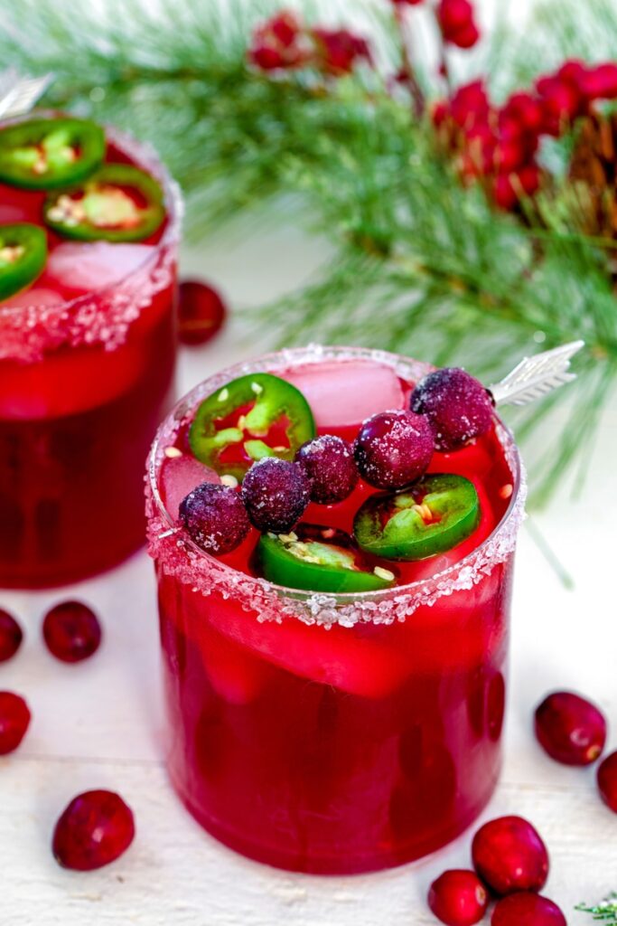 View of a cranberry jalapeño margarita with sugared cranberries and jalapeño round garnish with second cocktail and holiday holly in background