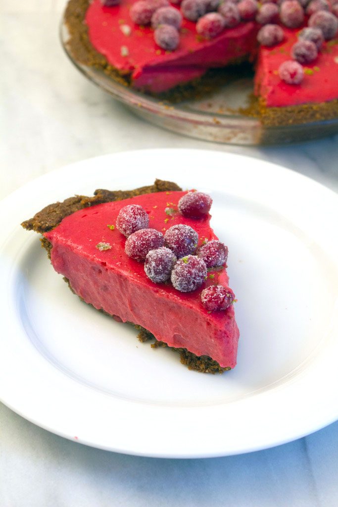 Head-on view of a slice of cranberry lime pie topped with sugared cranberries and lime zest on a white plate with remaining pie in the background