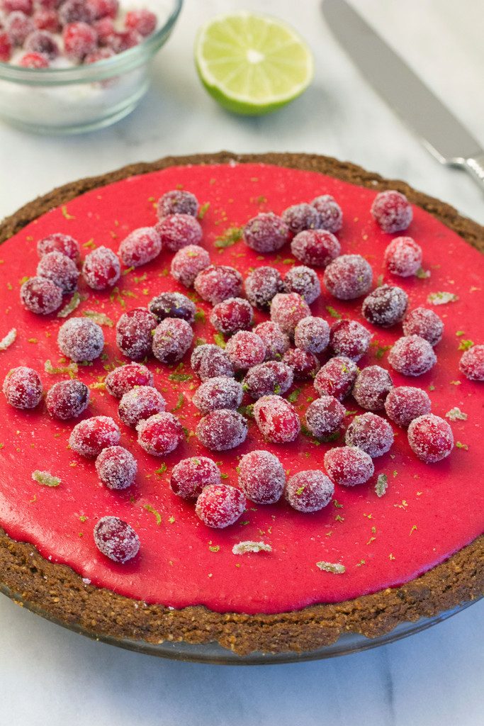 Head-on view of cranberry lime pie  topped with sugared cranberries and lime zest with knife, lime half, and bowl of sugared cranberries in background