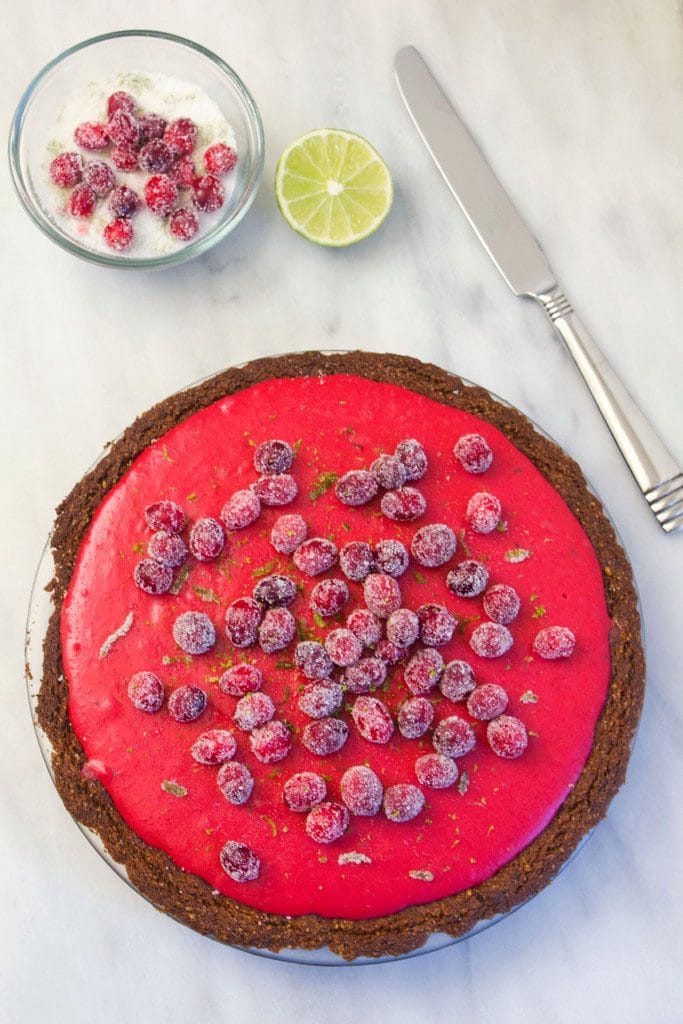 Overhead view of cranberry-lime pie topped with sugared cranberries and lime zest with a bowl of sugared cranberries, lime half, and knife in the background