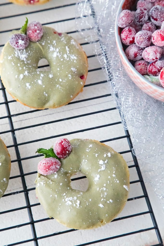 Overhead view of two cranberry matcha donuts on a baking rack with sugared cranberry garnish and bowl of sugared cranberries in the background