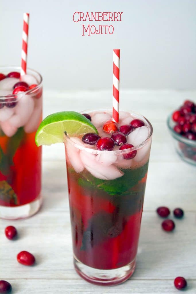 Head on view of cranberry mojito in tall glass with cranberry, mint, and lime garnish and red and white striped straw with second cocktail and cranberries in background and "Cranberry Mojito" text at top