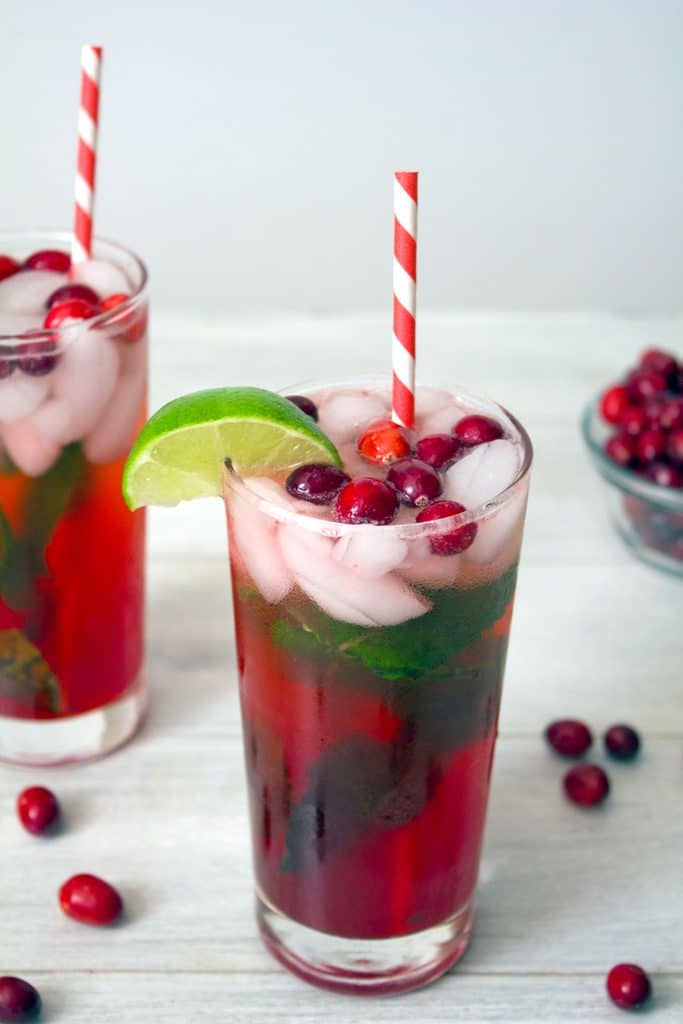 Head on view of cranberry mojito in tall glass with cranberry, mint, and lime garnish and red and white striped straw with second cocktail and cranberries in background